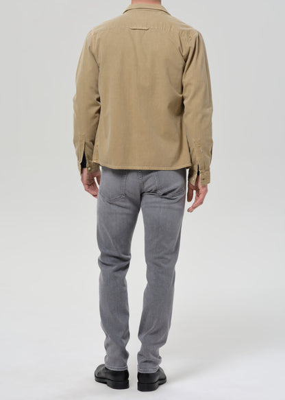 A man viewed from the back wearing a tan long-sleeve button-up shirt and grey slim-fit jeans, paired with black shoes, against a grey background.