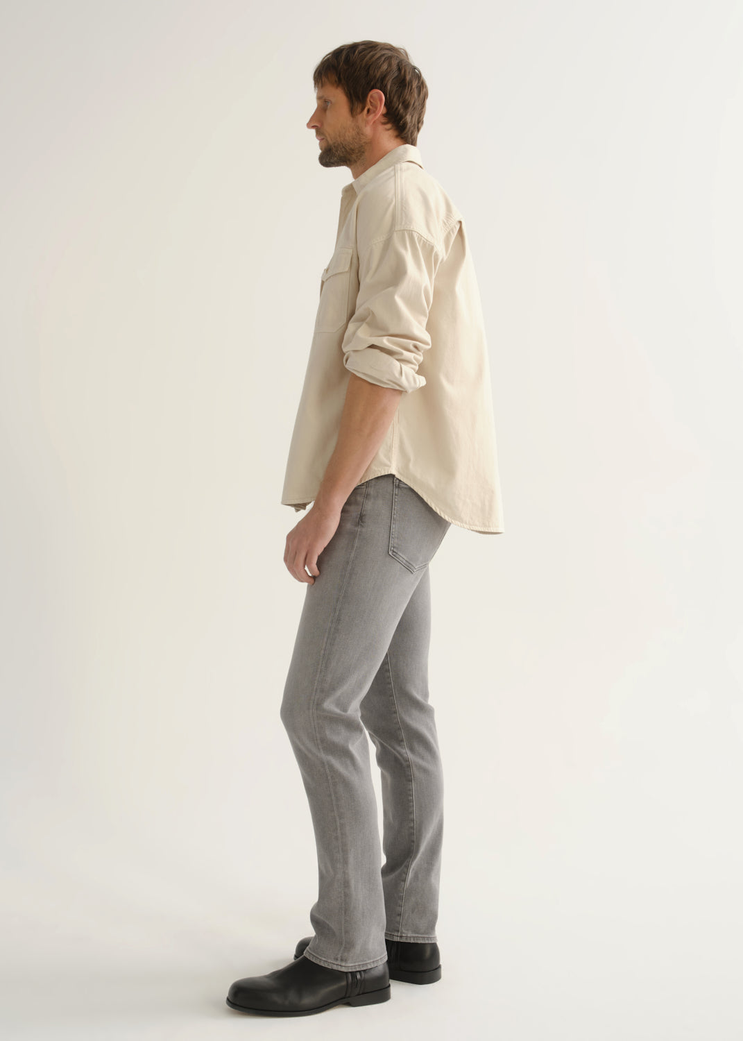 Person wearing a cream button-front shirt, grey jeans, and black boots, side full body view on a light grey background.