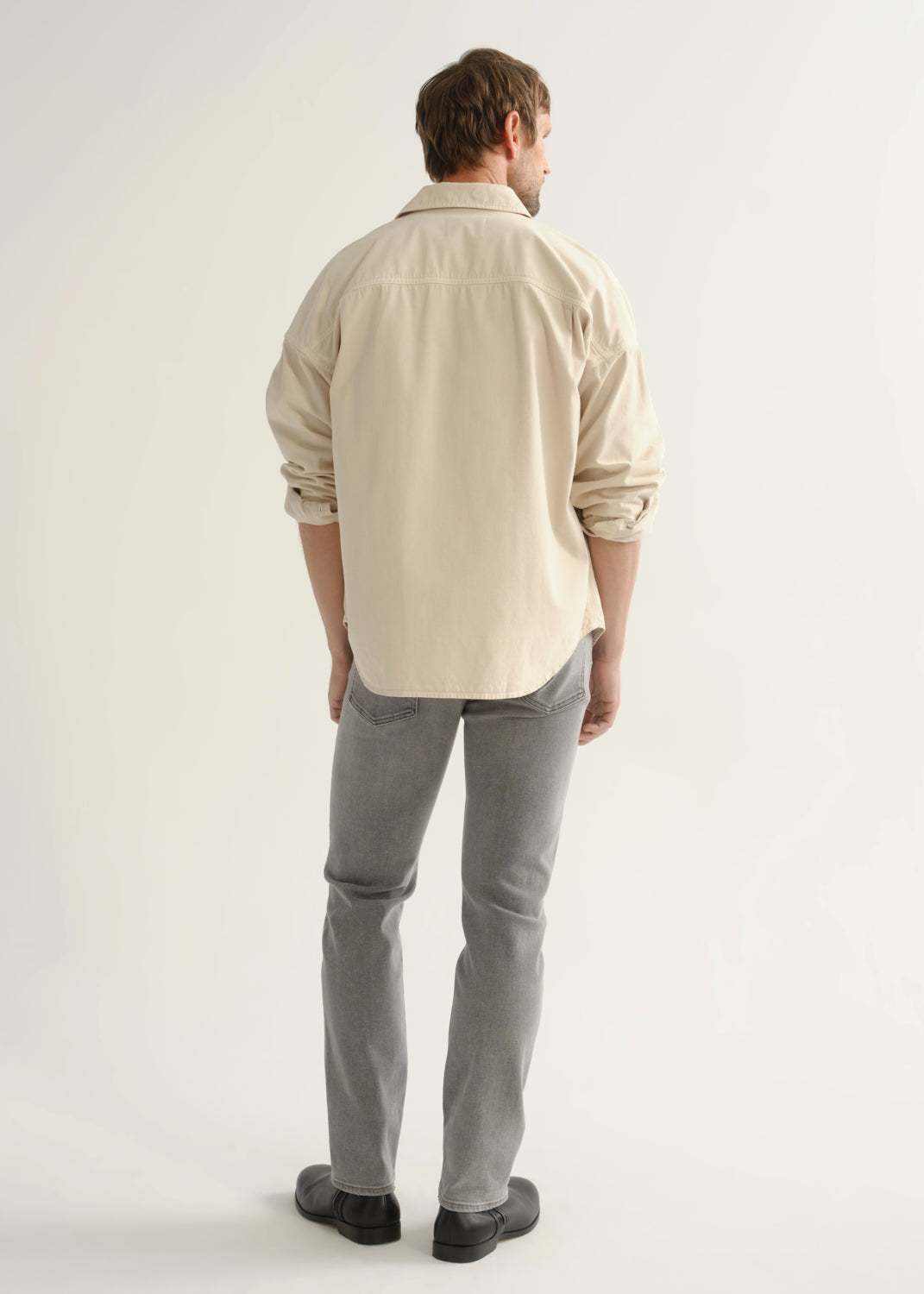 Person wearing a cream button-front shirt, grey jeans, and black boots, back full body view on a light grey background.