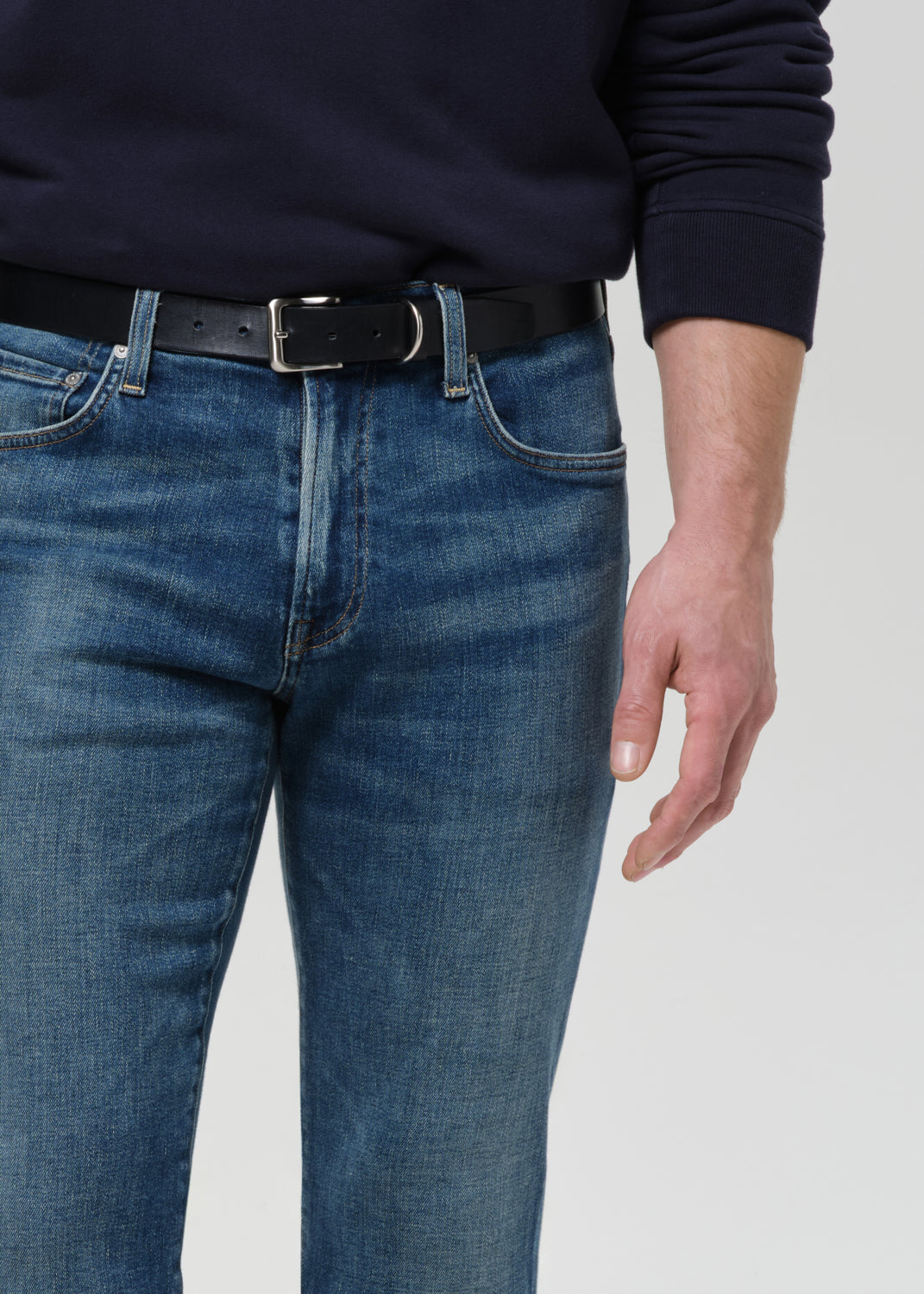 A close-up of a person wearing blue denim jeans with a black belt and a dark navy long-sleeve top, shown from the chest down against a grey background.