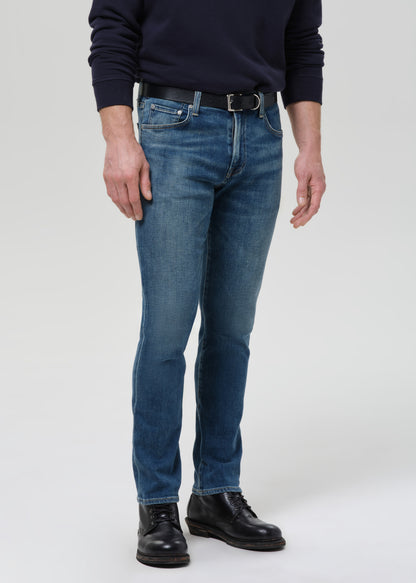 A person wearing blue slim-fit jeans with a black belt, a dark navy long-sleeve top, and black leather shoes, photographed against a grey background.