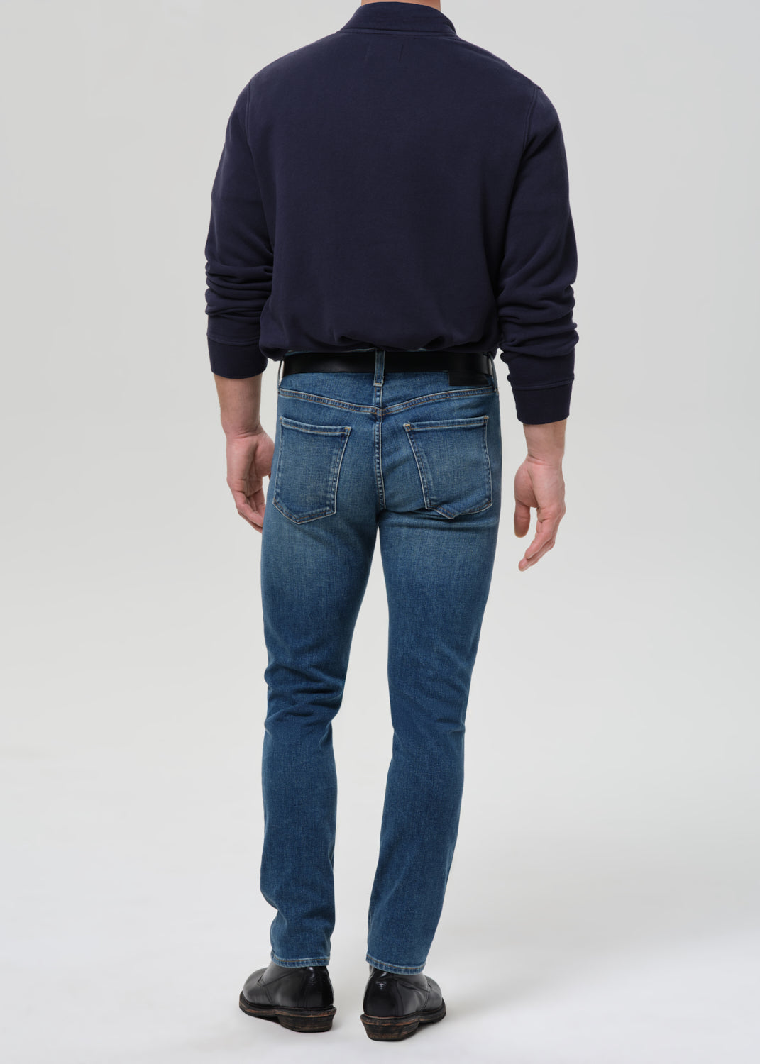 A man viewed from behind wearing a navy blue long-sleeve shirt, blue jeans, and dark brown leather shoes, standing against a grey background.