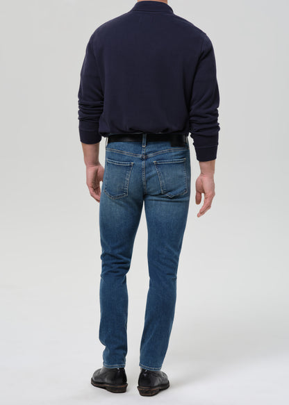 A man viewed from behind wearing a navy blue long-sleeve shirt, blue jeans, and dark brown leather shoes, standing against a grey background.