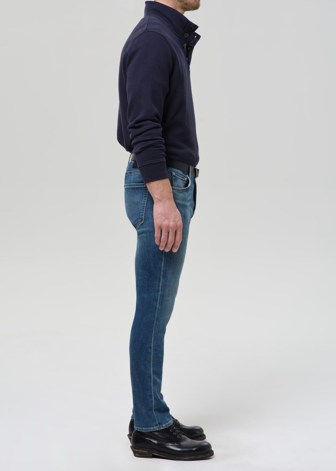 A man shown in profile wearing a dark navy long-sleeve quarter-zip pullover, blue jeans, and black leather shoes, standing against a grey background.