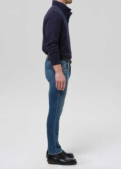 A man shown in profile wearing a dark navy long-sleeve quarter-zip pullover, blue jeans, and black leather shoes, standing against a grey background.