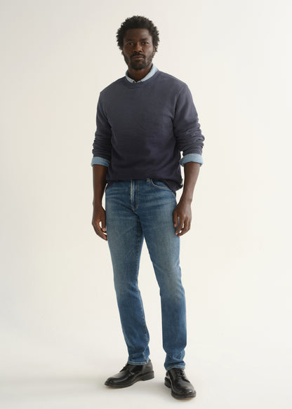 Person wearing medium blue straight leg jeans with a slate blue long sleeve collared pullover and black shoes, front full body view on a light grey background.