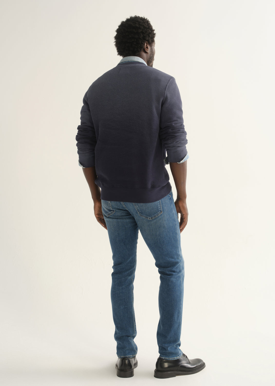 Person wearing medium blue straight leg jeans with a slate blue long sleeve collared pullover and black shoes, back full body view on a light grey background.