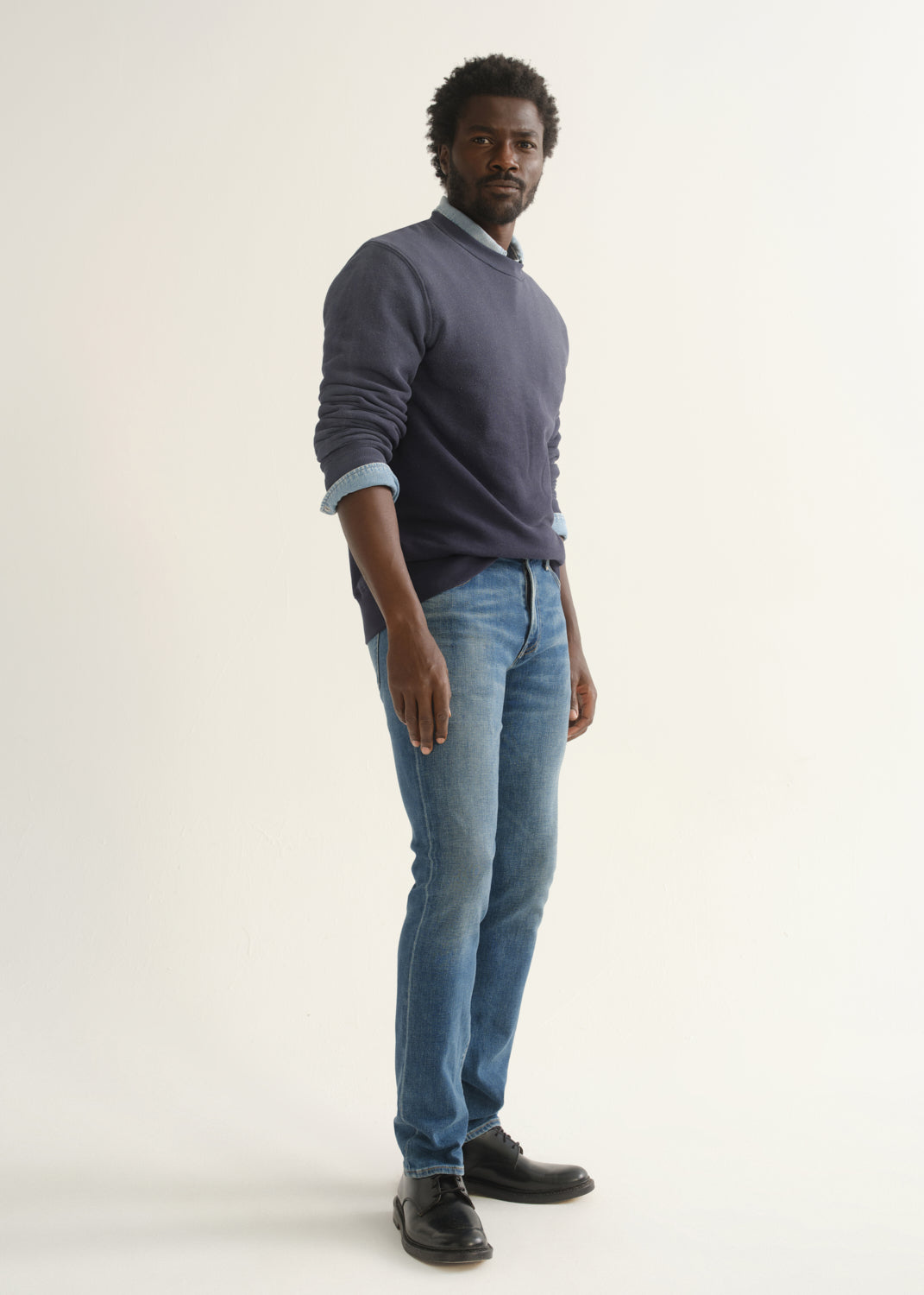 Person wearing medium blue straight leg jeans with a slate blue long sleeve collared pullover and black shoes, angled front full body view on a light grey background.