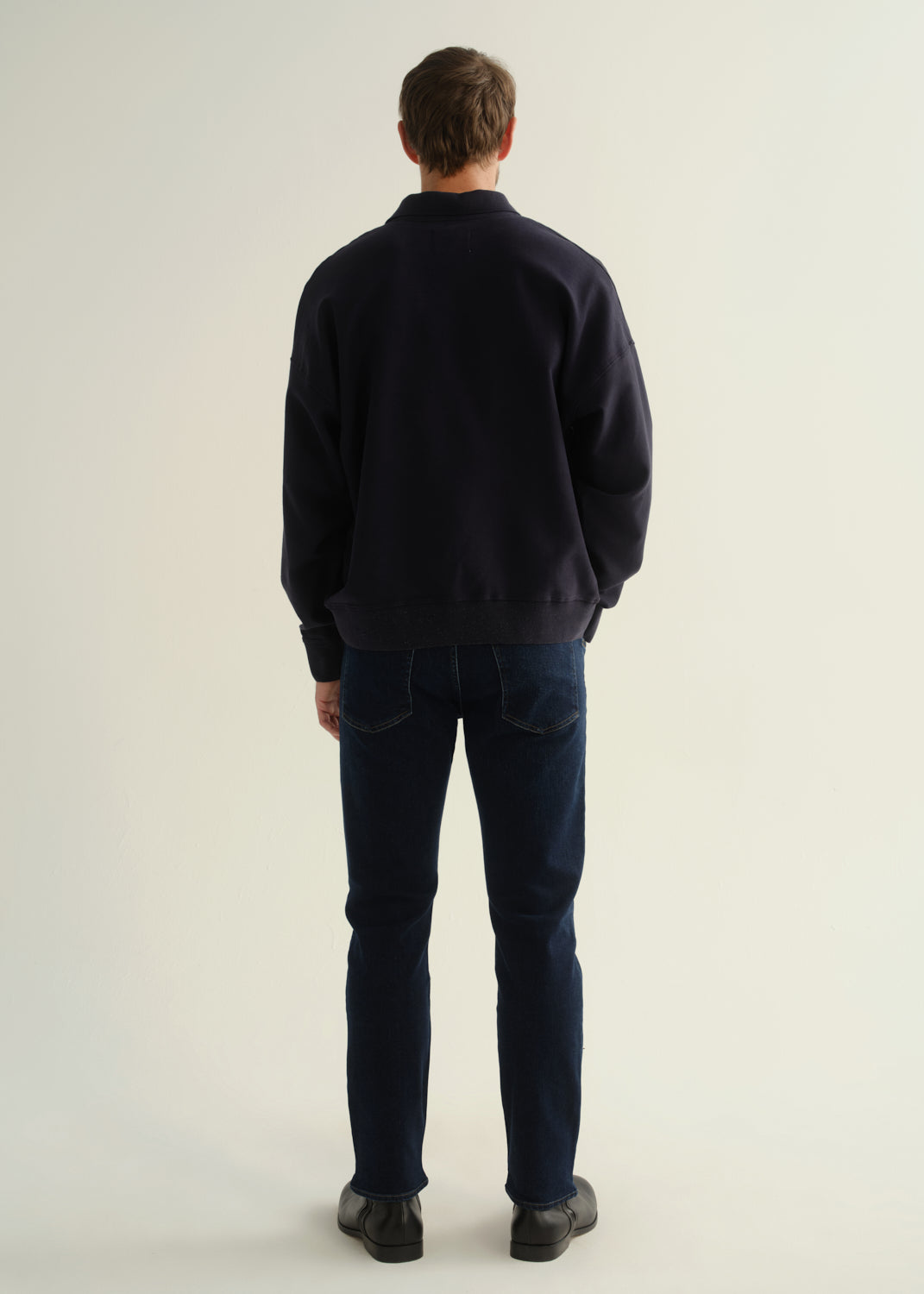Person wearing a dark navy long sleeve shirt, dark indigo jeans, and black boots, back full body view on a light grey background.