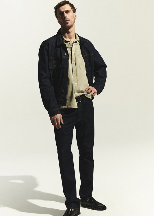 Man wearing a dark denim jacket, beige shirt, and dark wash jeans on a white background