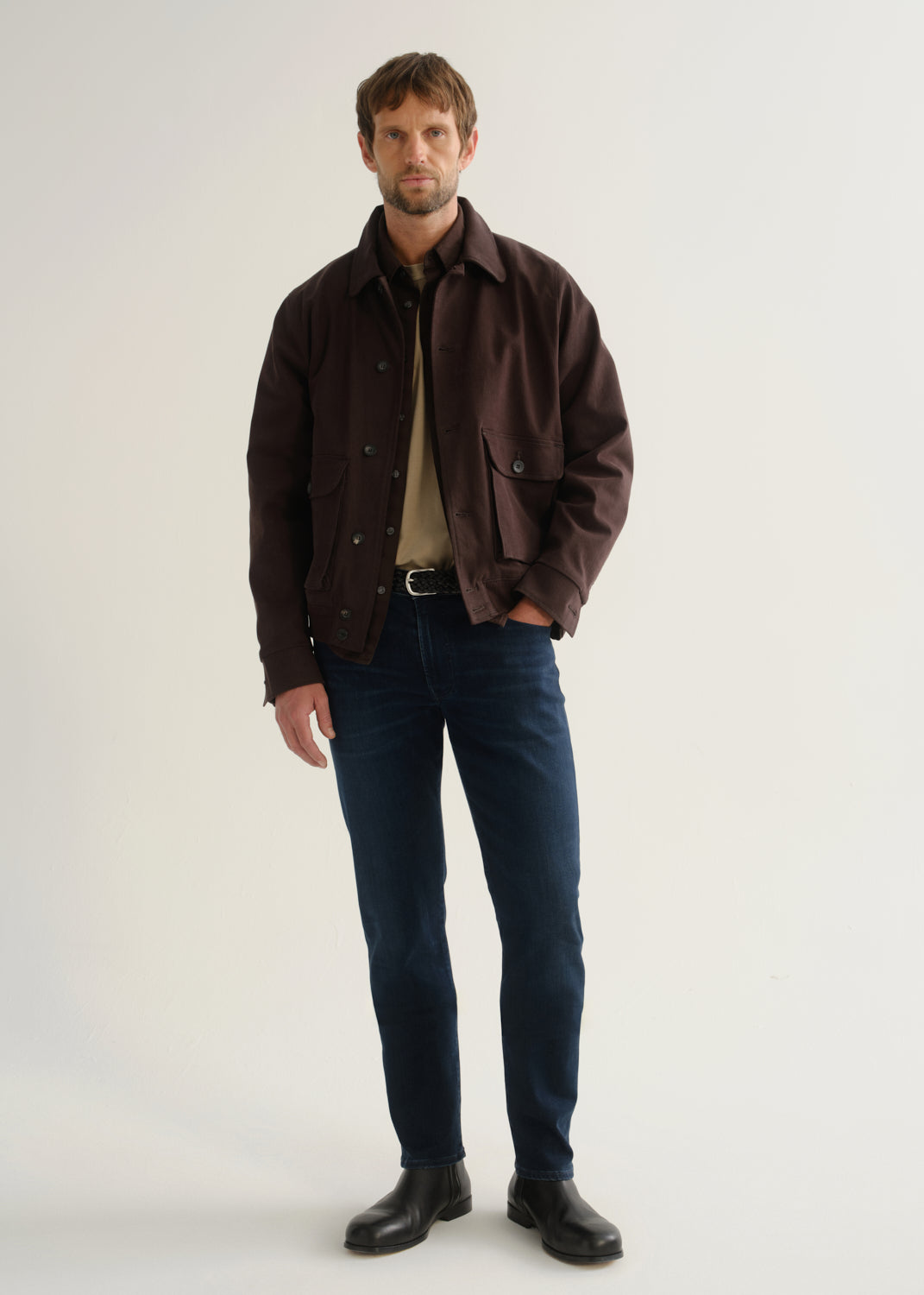 Person wearing dark indigo blue jeans, a dark brown jacket, a black shirt, and black boots, front full body view on a light grey background.