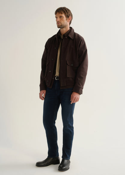 Person wearing dark indigo blue jeans, a dark brown jacket, a black shirt, and black boots, angled front full body view on a light grey background.