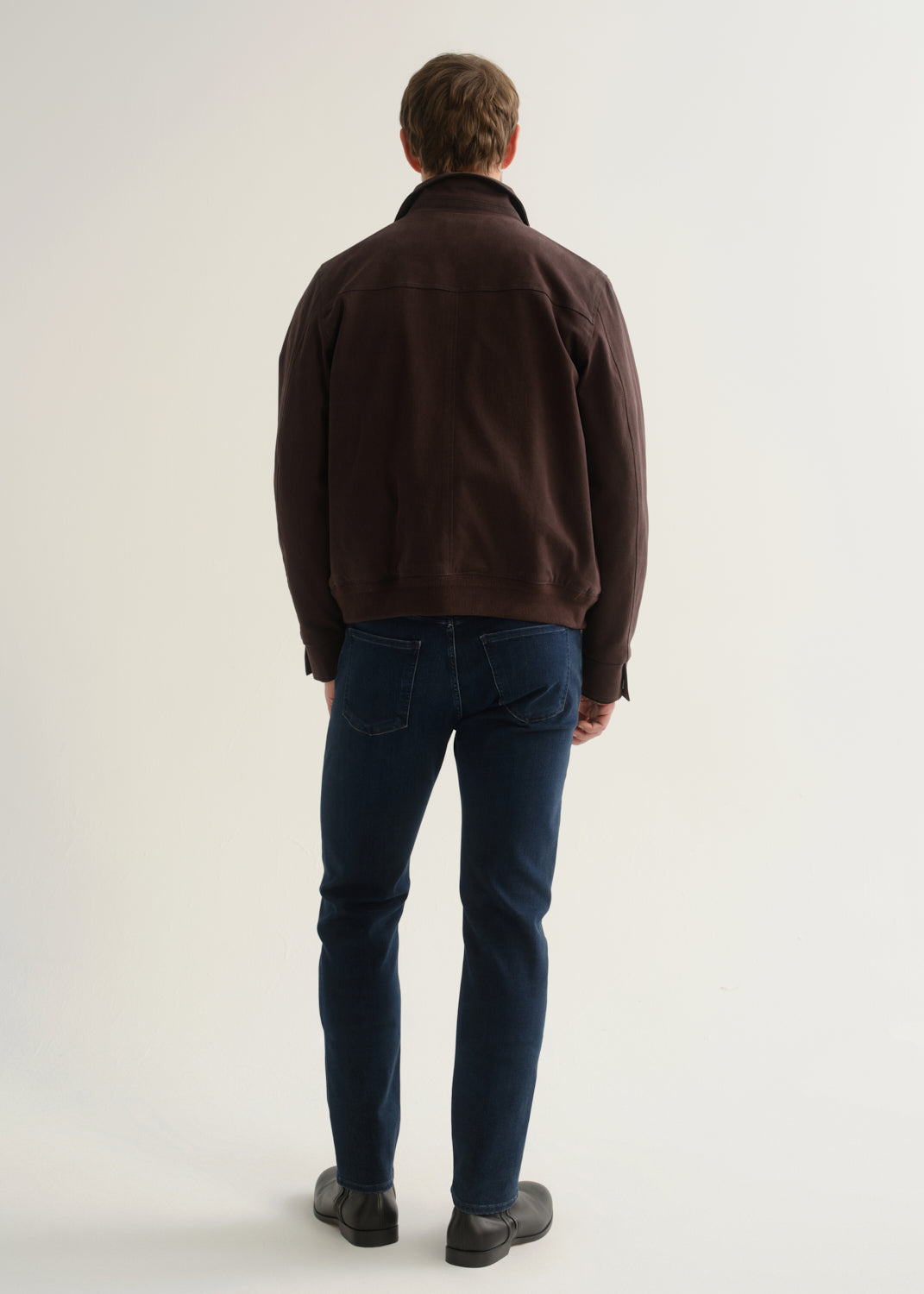 Person wearing dark indigo blue jeans, a dark brown jacket, and black boots, back full body view on a light grey background.