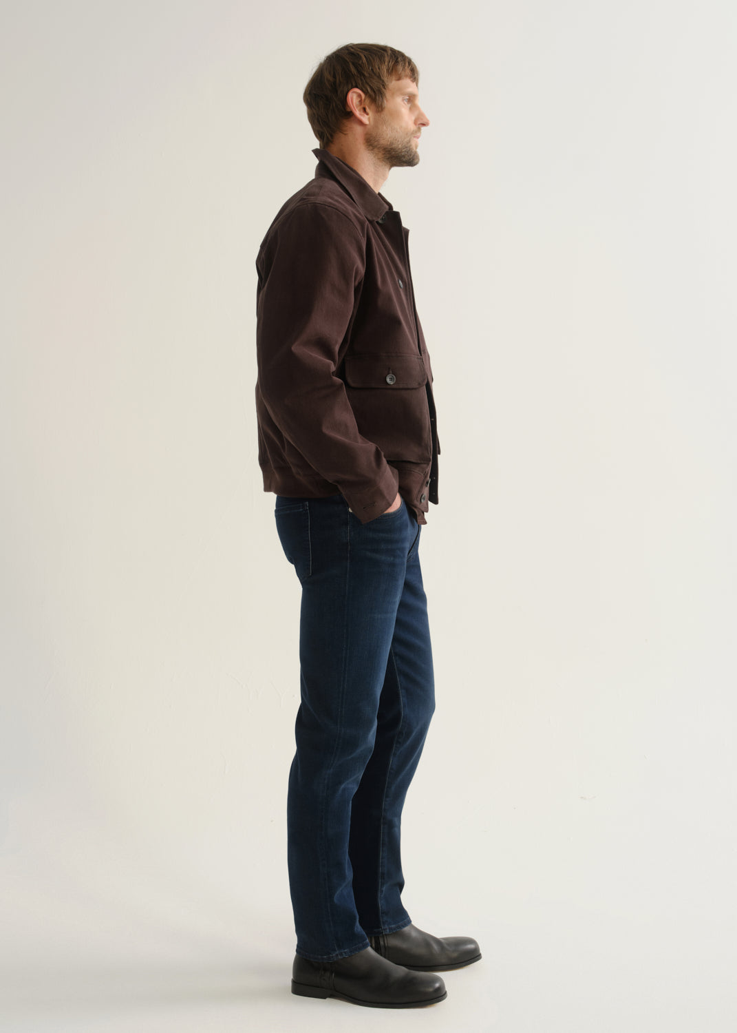 Person wearing dark indigo blue jeans, a dark brown jacket, and black boots, side full body view on a light grey background.