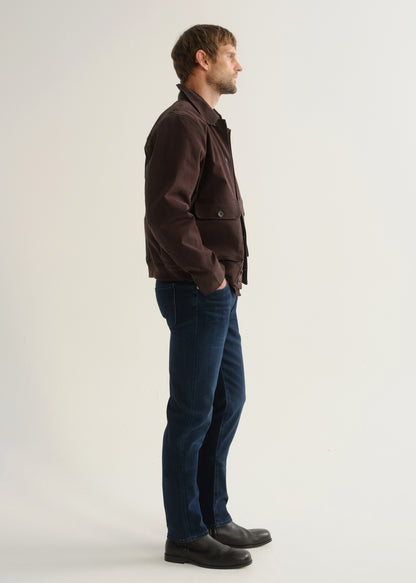 Person wearing dark indigo blue jeans, a dark brown jacket, and black boots, side full body view on a light grey background.