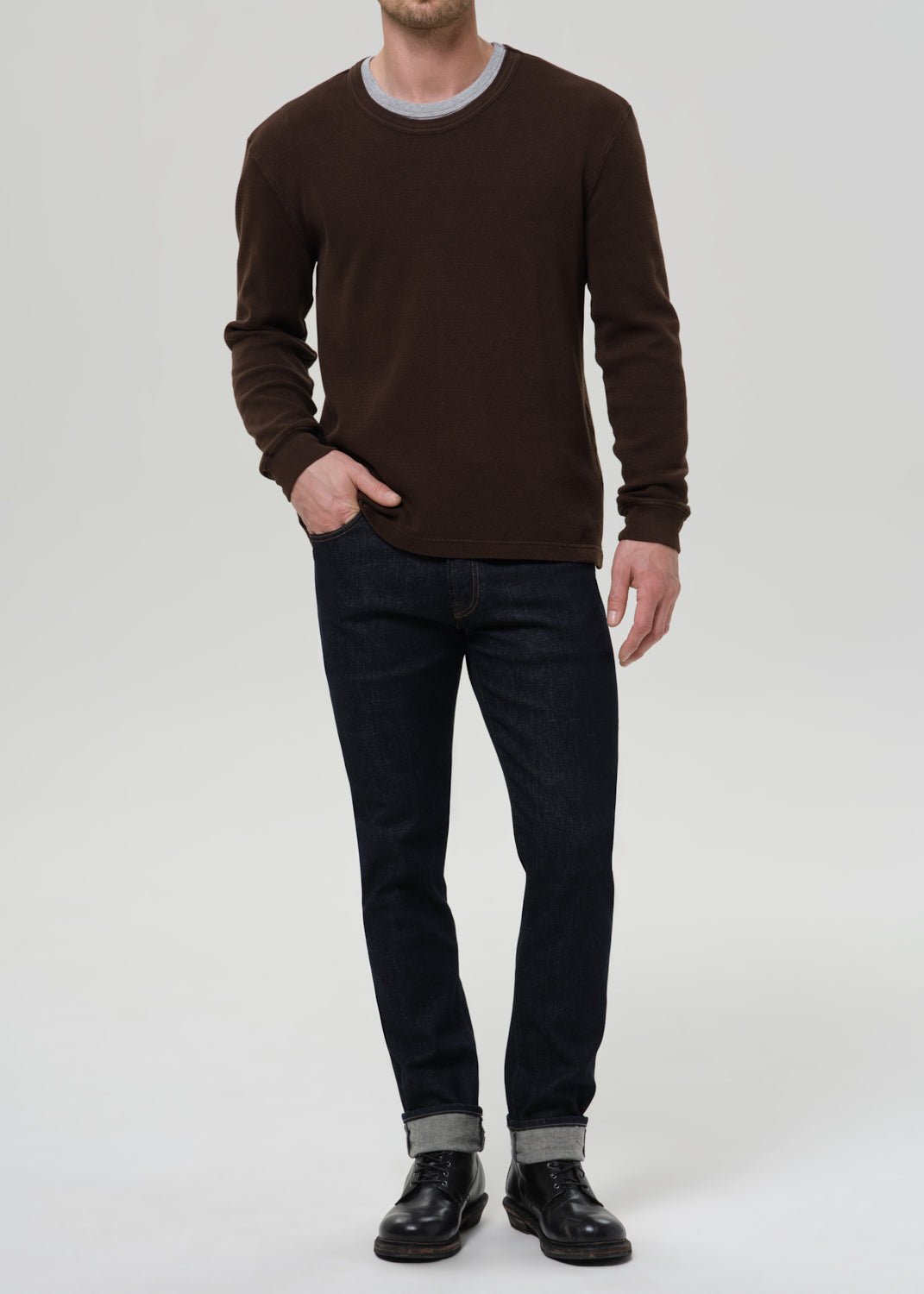 Person wearing dark indigo straight leg jeans with a brown crewneck sweater on a grey background.