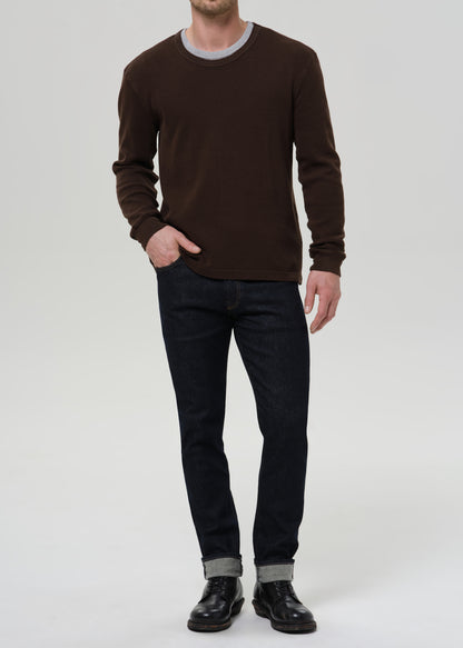 Person wearing dark indigo straight leg jeans with a brown crewneck sweater on a grey background.