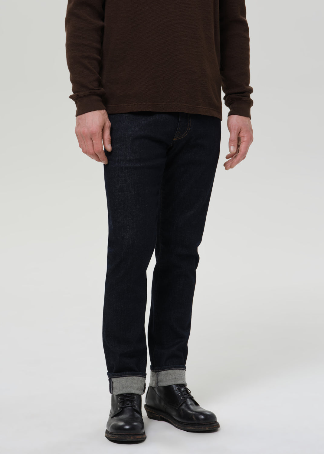 Person wearing dark indigo straight leg jeans with a brown crewneck sweater on a grey background.