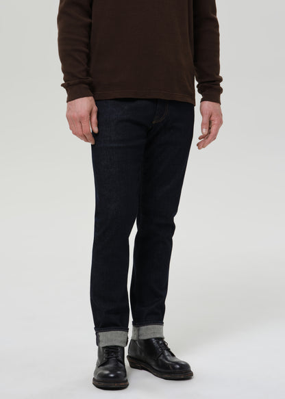 Person wearing dark indigo straight leg jeans with a brown crewneck sweater on a grey background.