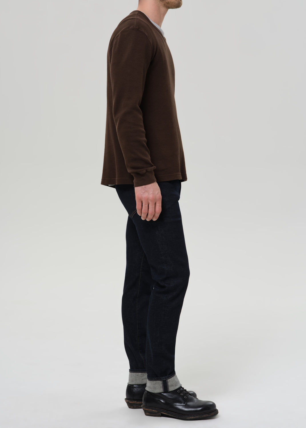 Person wearing dark indigo straight leg jeans with a brown crewneck sweater viewed from the side on a grey background.