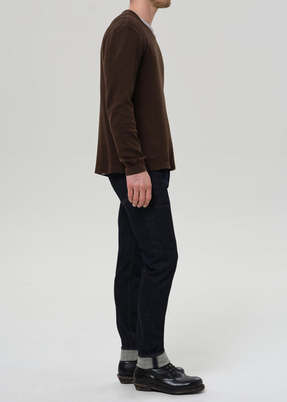 Person wearing dark indigo straight leg jeans with a brown crewneck sweater viewed from the side on a grey background.