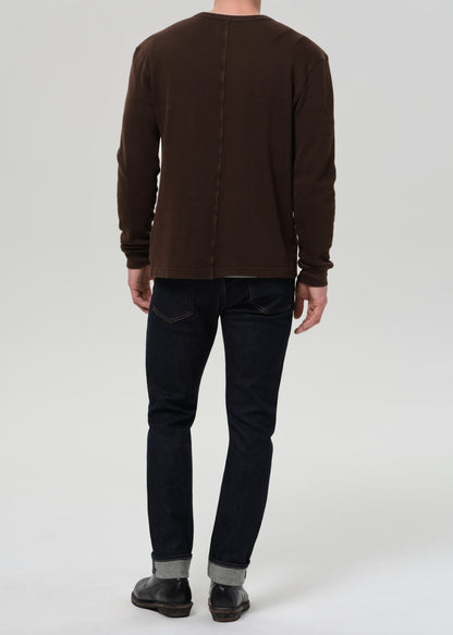 Person wearing dark indigo straight leg jeans with a brown crewneck sweater viewed from the back on a grey background.