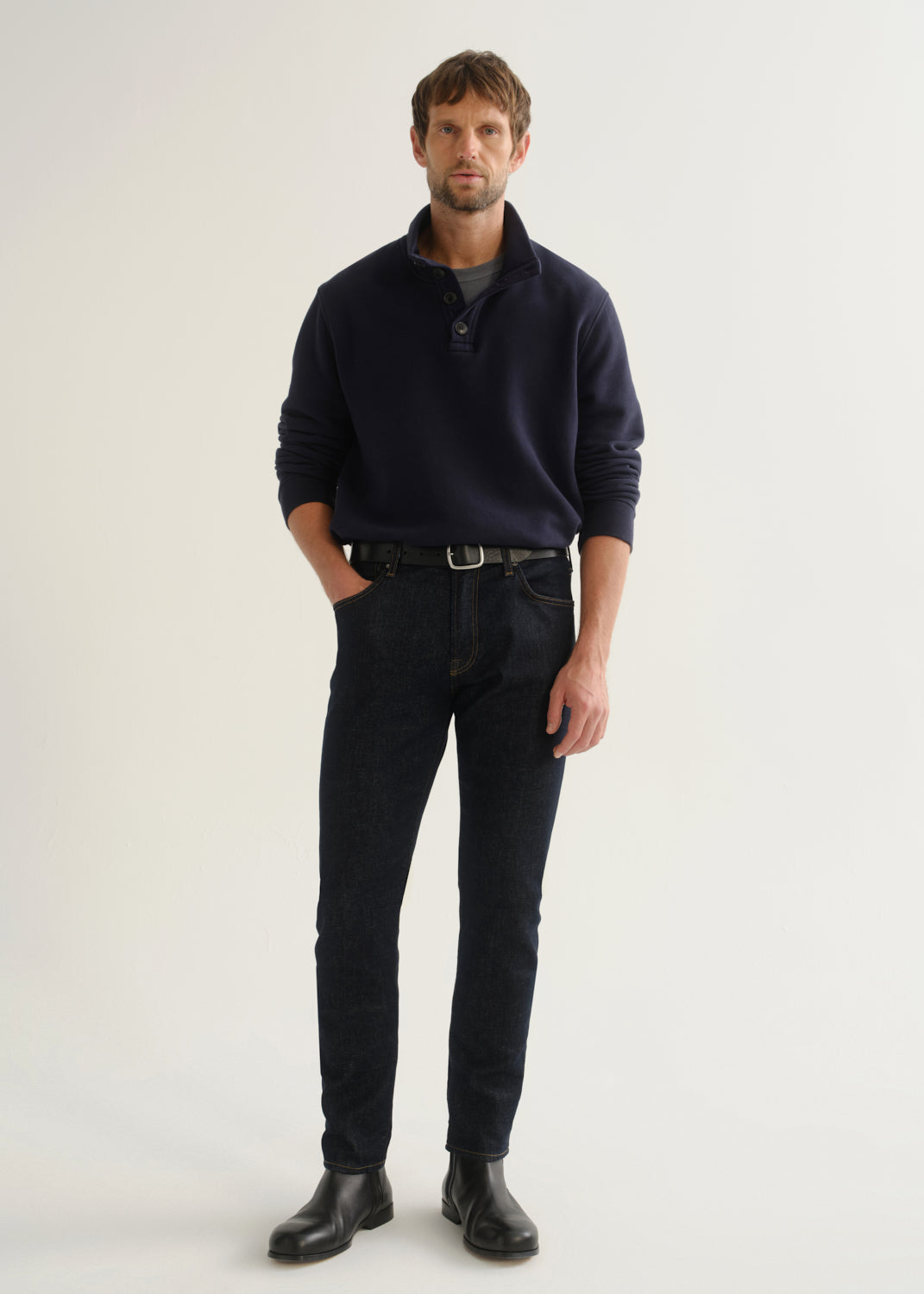 Person wearing dark indigo blue jeans, a navy long sleeve shirt, and black boots, front full body view on a light grey background.
