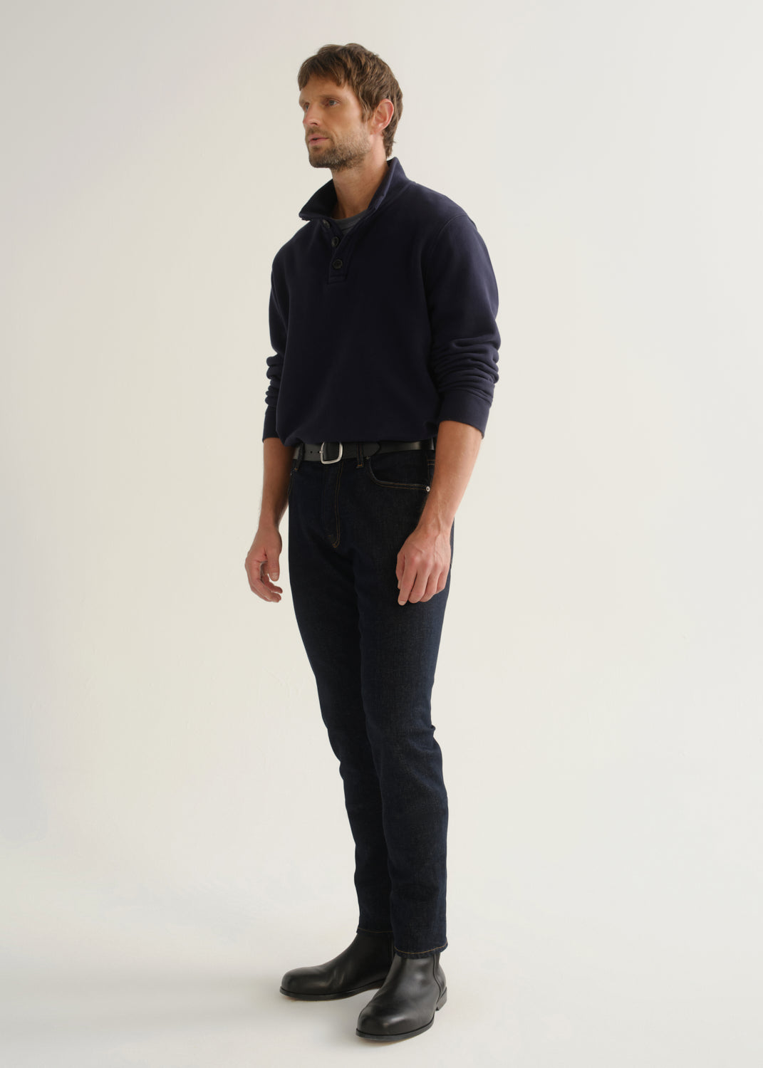 Person wearing dark indigo blue jeans, a navy long sleeve shirt, and black boots, angled front full body view on a light grey background.