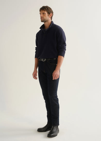 Person wearing dark indigo blue jeans, a navy long sleeve shirt, and black boots, angled front full body view on a light grey background.