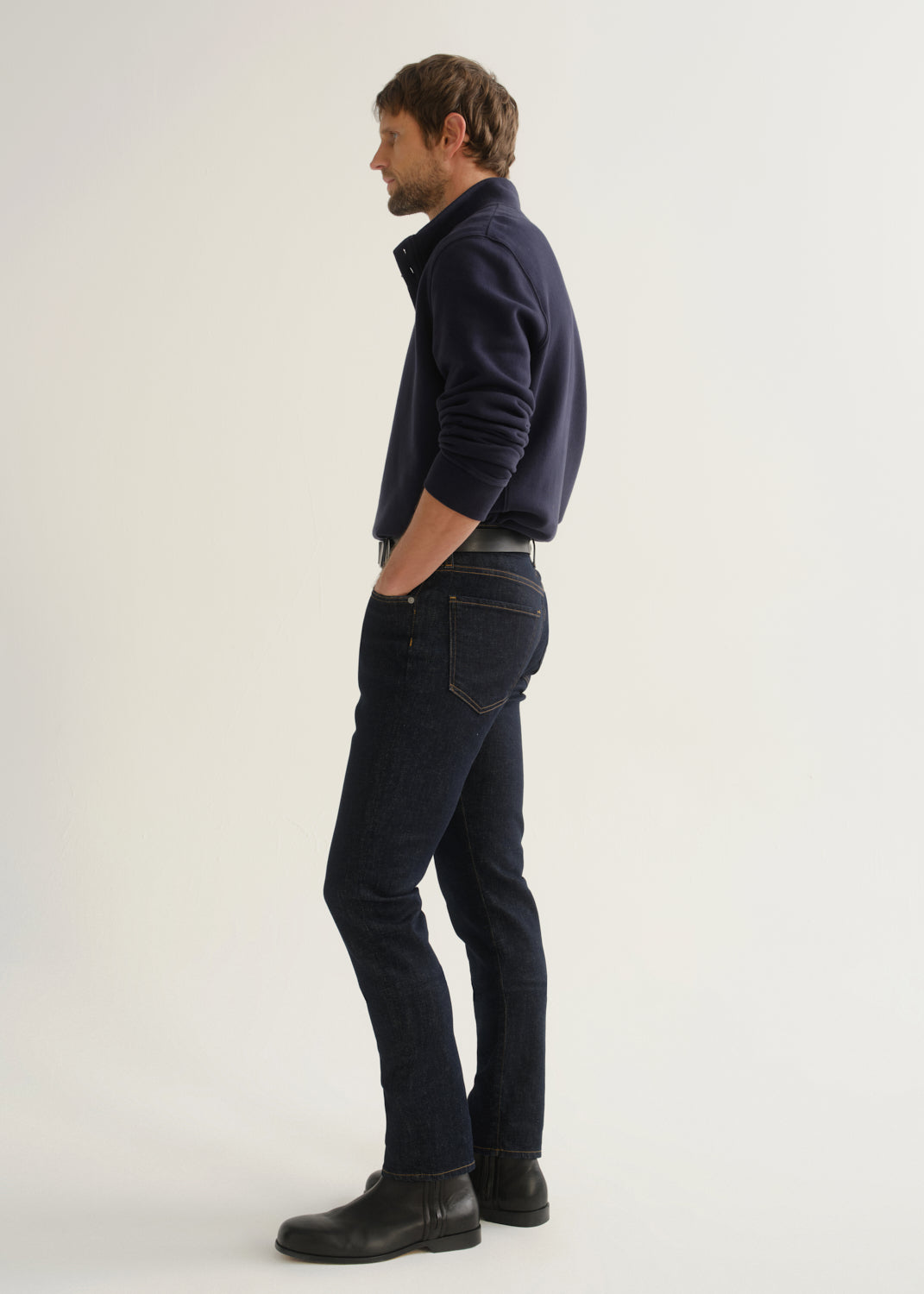 Person wearing dark indigo blue jeans, a navy long sleeve shirt, and black boots, side full body view on a light grey background.