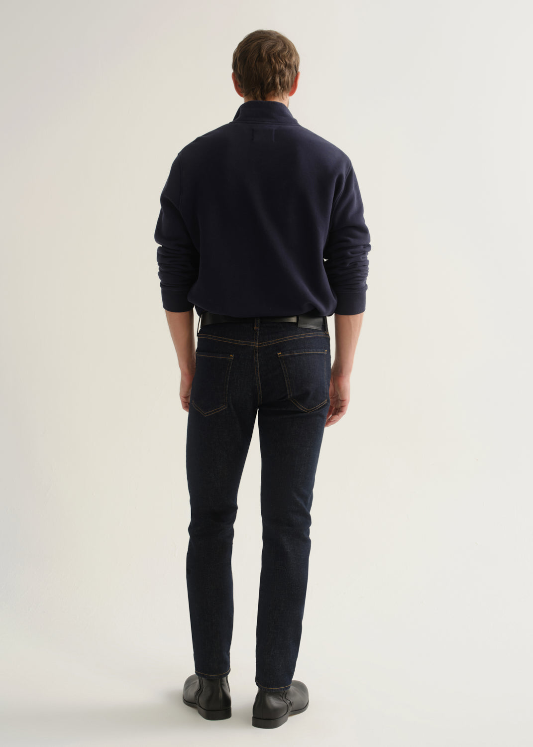 Person wearing dark indigo blue jeans, a navy long sleeve shirt, and black boots, back full body view on a light grey background.