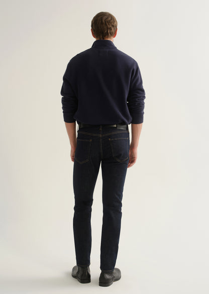 Person wearing dark indigo blue jeans, a navy long sleeve shirt, and black boots, back full body view on a light grey background.
