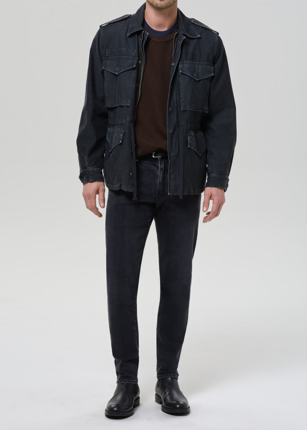 Person wearing dark indigo straight leg jeans with a black denim jacket and brown crewneck tee on a grey background.