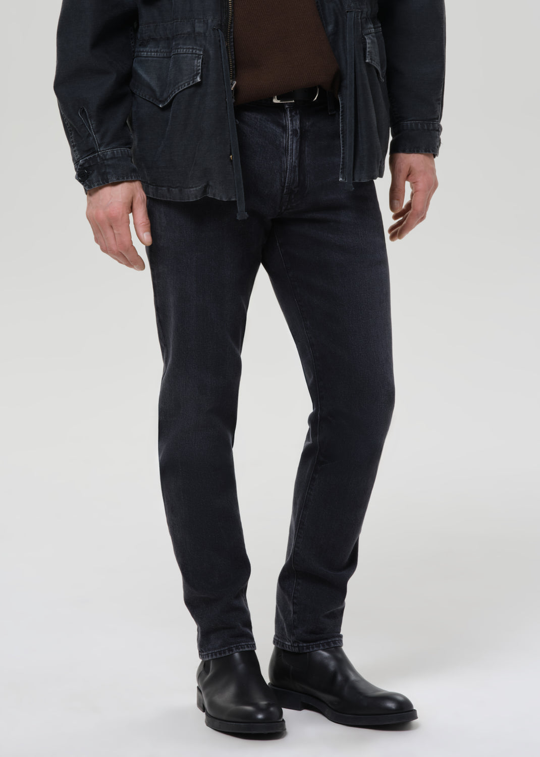 Person wearing dark indigo straight leg jeans with a black denim jacket and brown crewneck tee on a grey background.