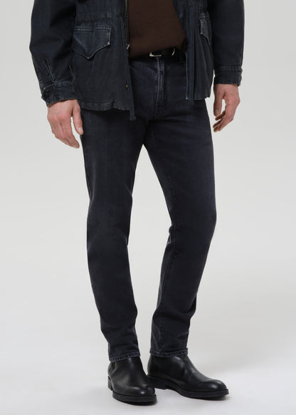 Person wearing dark indigo straight leg jeans with a black denim jacket and brown crewneck tee on a grey background.