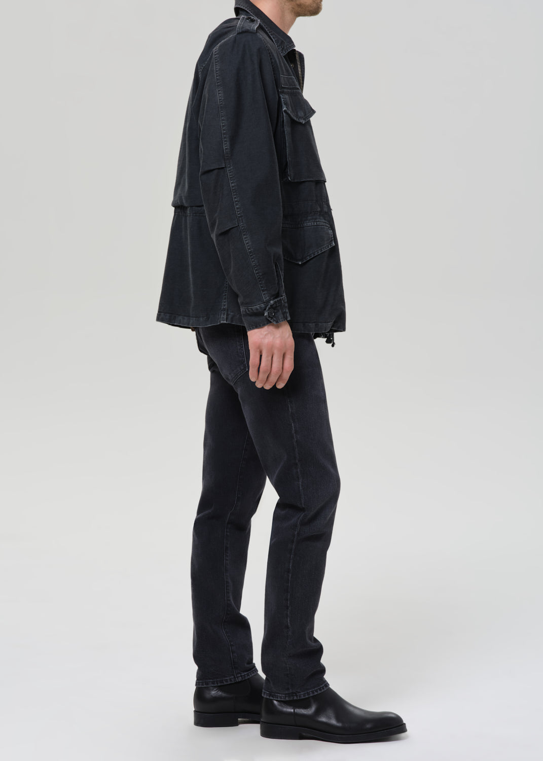Person wearing dark indigo straight leg jeans with a black denim jacket and brown crewneck tee viewed from the side on a grey background.
