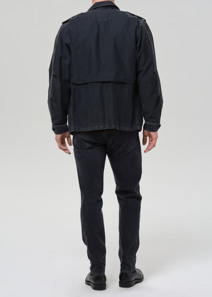 Person wearing dark indigo straight leg jeans with a black denim jacket and brown crewneck tee viewed from the back on a grey background.