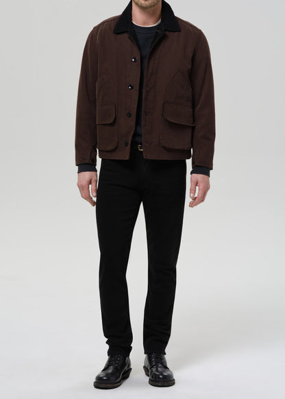 Person wearing black straight leg jeans with a brown chore jacket and black crewneck tee on a grey background.