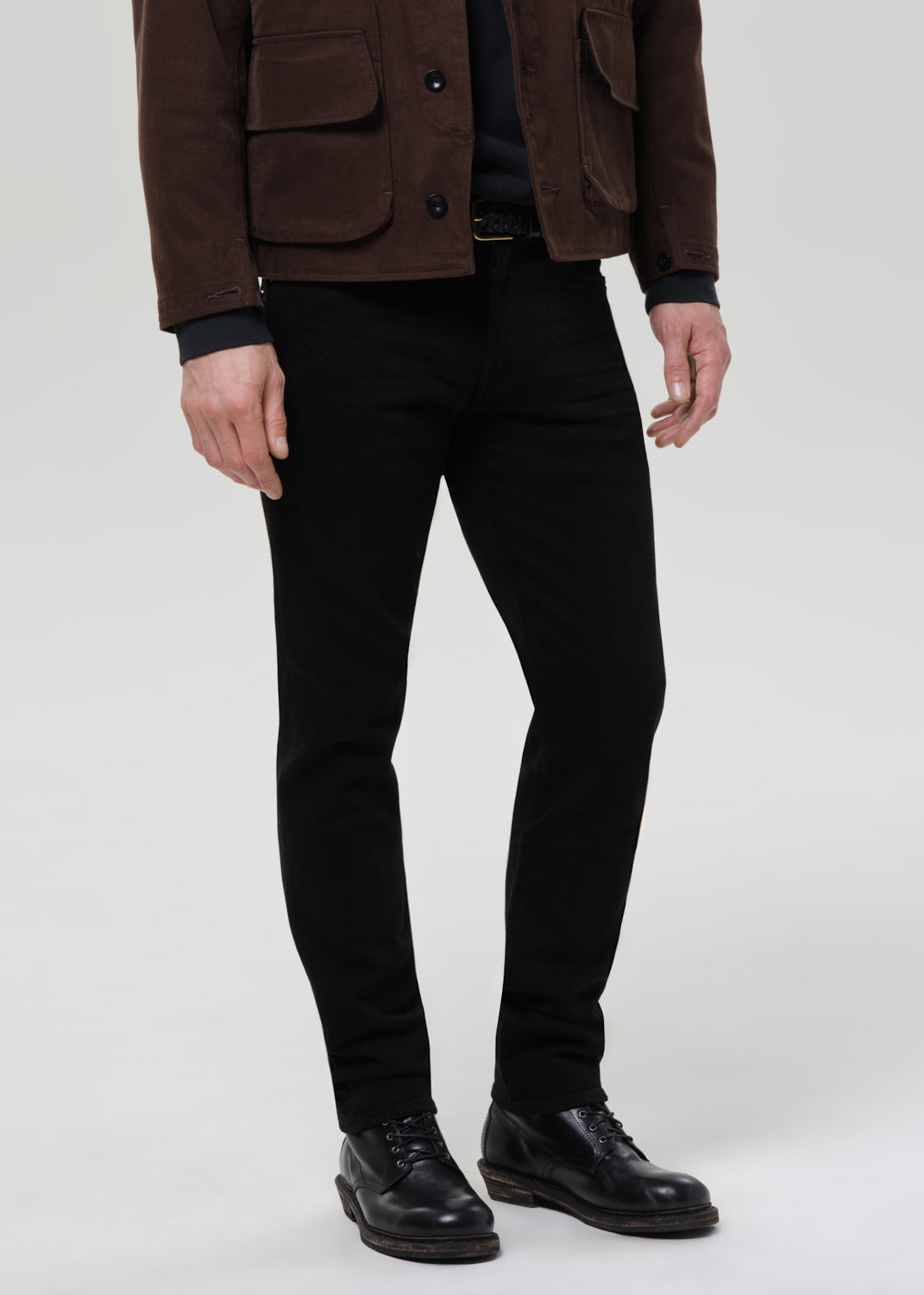 Person wearing black straight leg jeans with a brown chore jacket and black crewneck tee on a grey background.