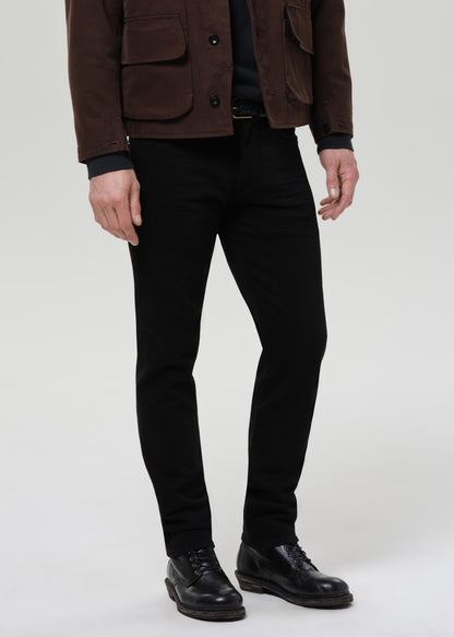 Person wearing black straight leg jeans with a brown chore jacket and black crewneck tee on a grey background.