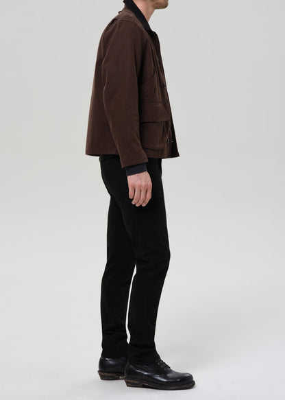 Person wearing black straight leg jeans with a brown chore jacket and black crewneck tee viewed from the side on a grey background.