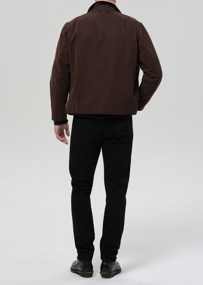 Person wearing black straight leg jeans with a brown chore jacket and black crewneck tee viewed from the back on a grey background.
