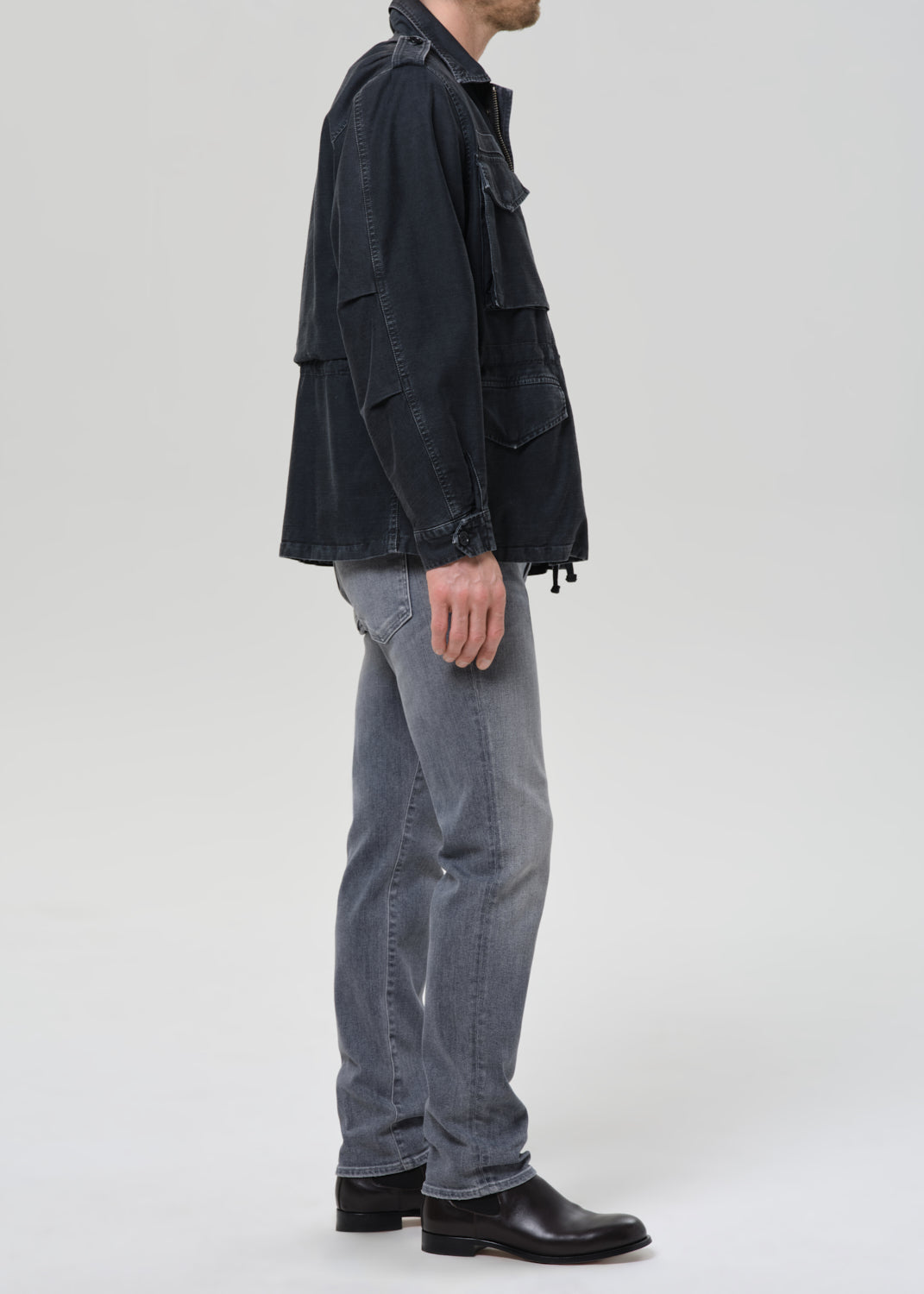 Person wearing light grey straight leg jeans with a black denim jacket and black crewneck tee viewed from the side on a grey background.