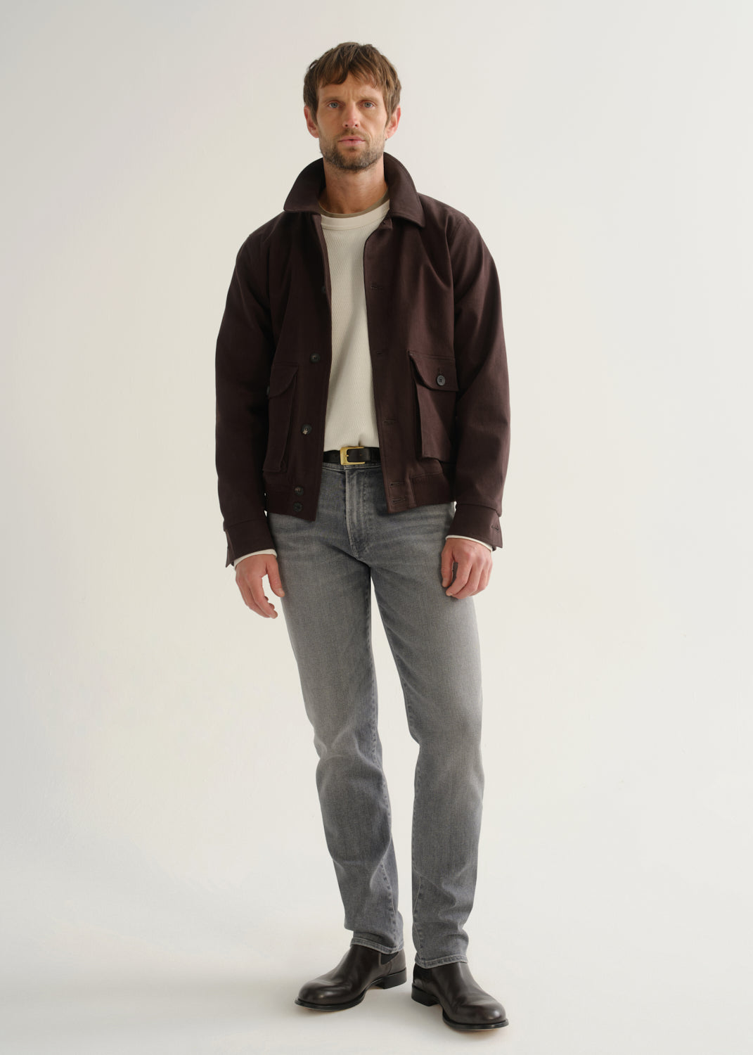 Person wearing washed grey jeans, a dark brown jacket, a white shirt, and black boots, front full body view on a light grey background.