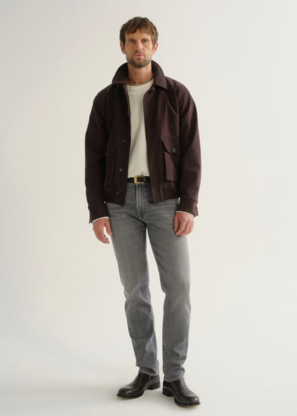 Person wearing washed grey jeans, a dark brown jacket, a white shirt, and black boots, front full body view on a light grey background.