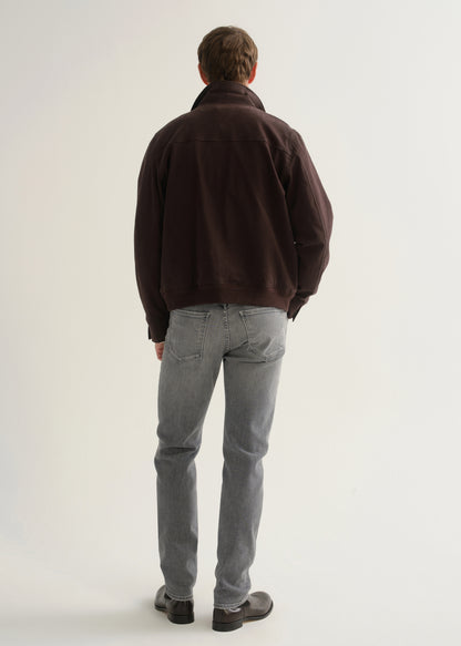 Person wearing washed grey jeans, a dark brown jacket, and black boots, back full body view on a light grey background.