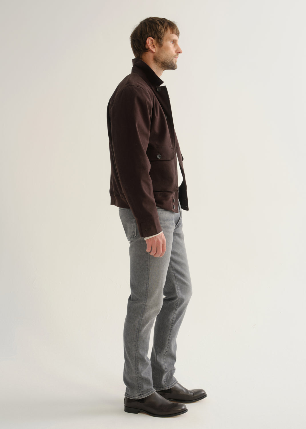 Person wearing washed grey jeans, a dark brown jacket, and black boots, side full body view on a light grey background.