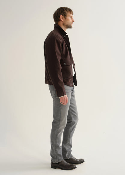 Person wearing washed grey jeans, a dark brown jacket, and black boots, side full body view on a light grey background.