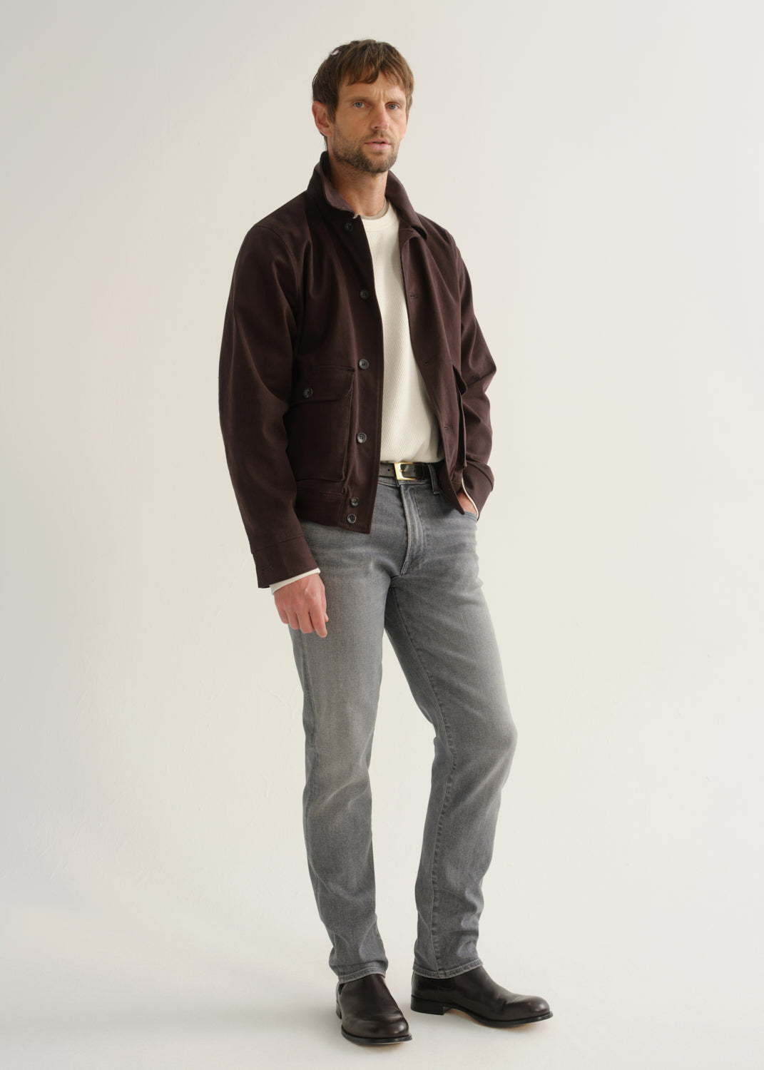 Person wearing washed grey jeans, a dark brown jacket, a white shirt, and black boots, angled front full body view on a light grey background.