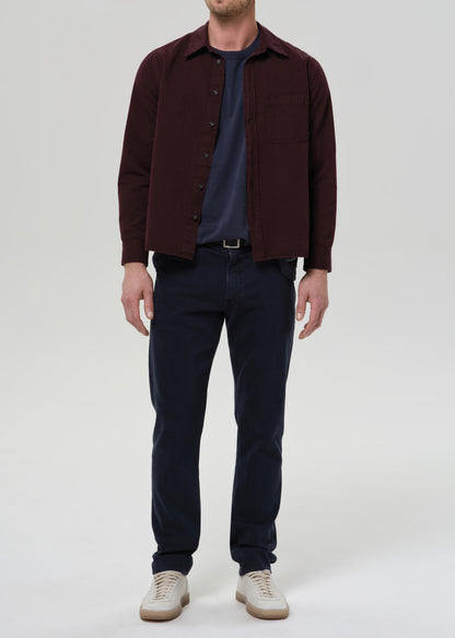 A man wearing a dark burgundy overshirt over a navy blue t-shirt, dark navy jeans, and off-white sneakers, standing against a grey background.